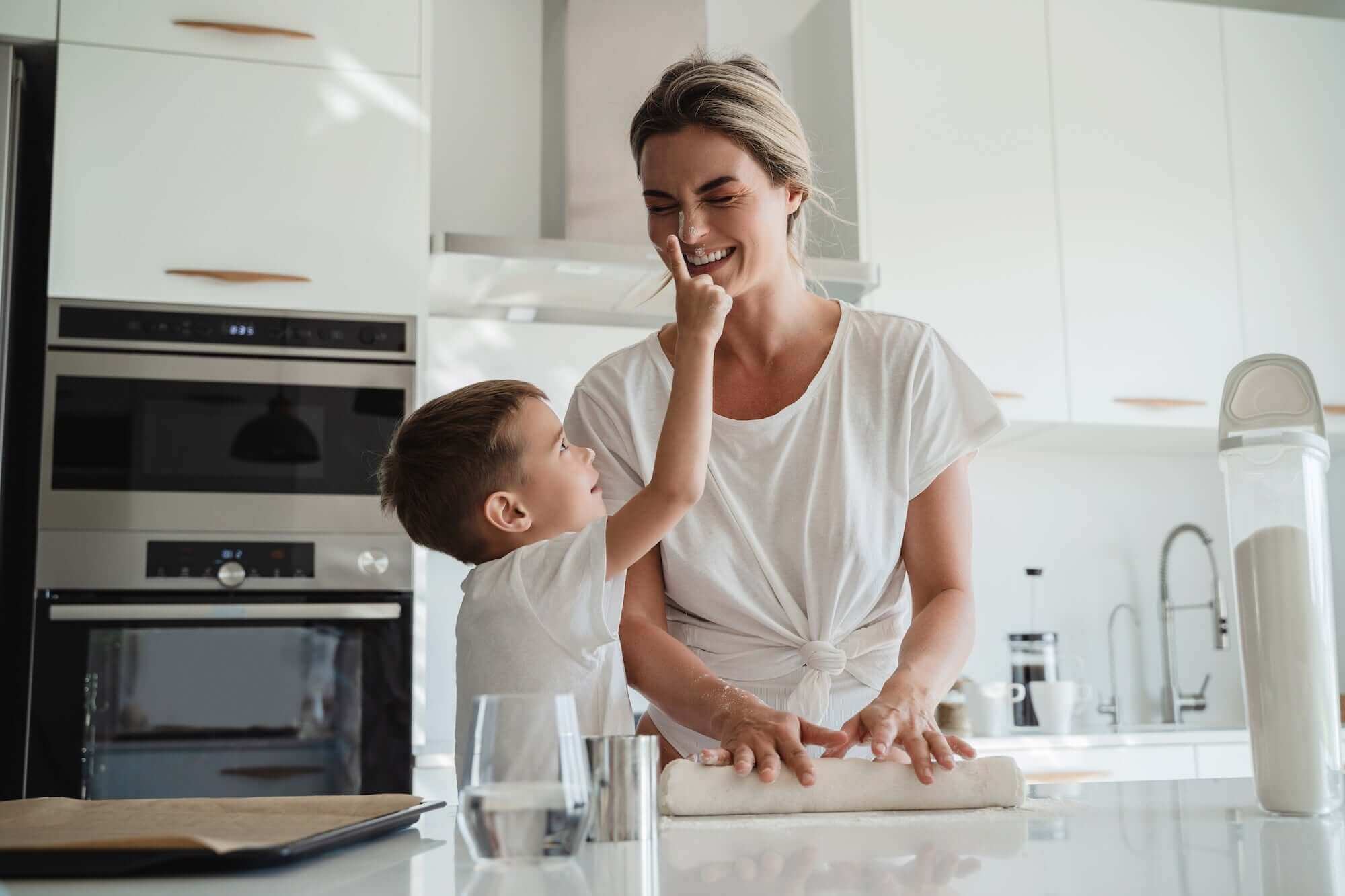 Mutter mit Sohn in einer weißen, modernen Küche, die gemeinsam Teig ausrollen.